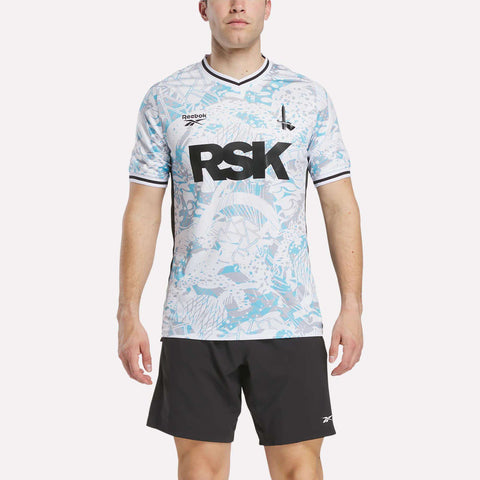 A man faces forward wearing the Men's Charlton Away Jersey by Reebok, featuring aquatic blue patterns, club heritage accents, bold black RSK lettering on the chest, and paired with black shorts.