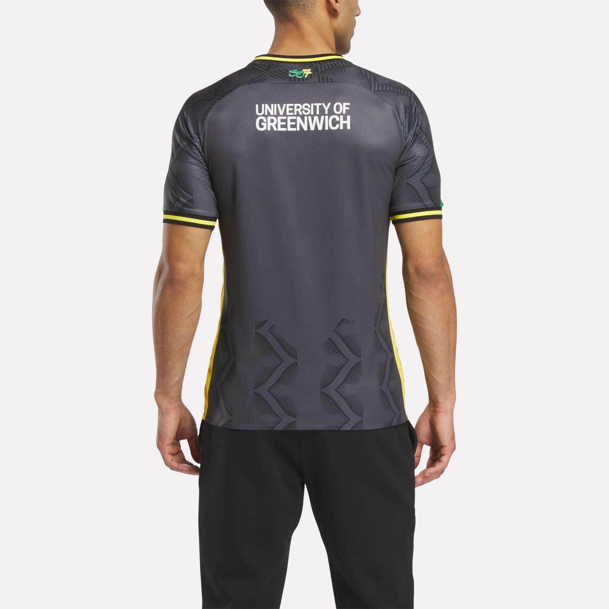A man faces away from the camera wearing the Reebok Men's Charlton Third Jersey, a dark athletic shirt with yellow accents, University of Greenwich on the upper back, and lightweight fabric. He pairs it with black pants.