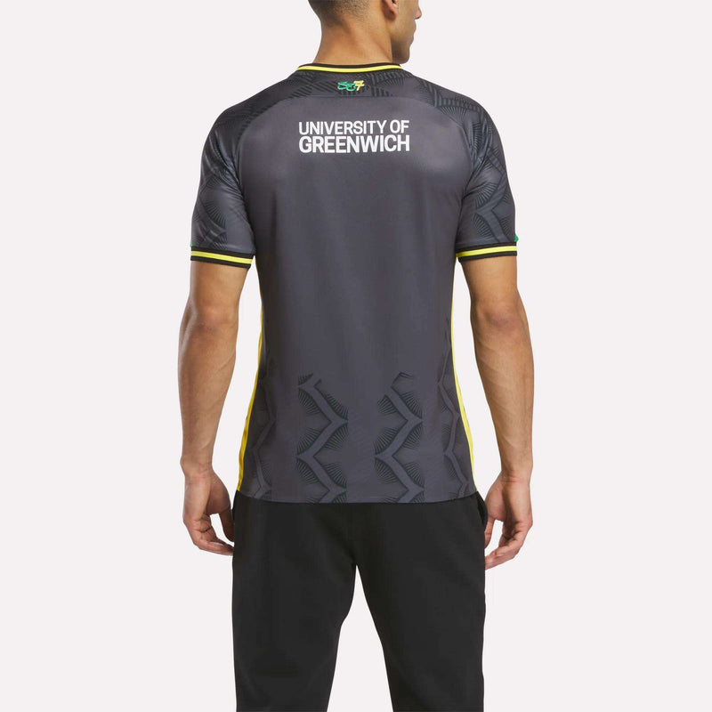 A man faces away from the camera wearing the Reebok Men's Charlton Third Jersey, a dark athletic shirt with yellow accents, University of Greenwich on the upper back, and lightweight fabric. He pairs it with black pants.