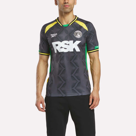 A man wears a Reebok Men's Charlton Third Jersey made of lightweight fabric in dark gray with yellow, green, and black accents. "RSK" appears in large white letters on the front as he stands against a plain background.