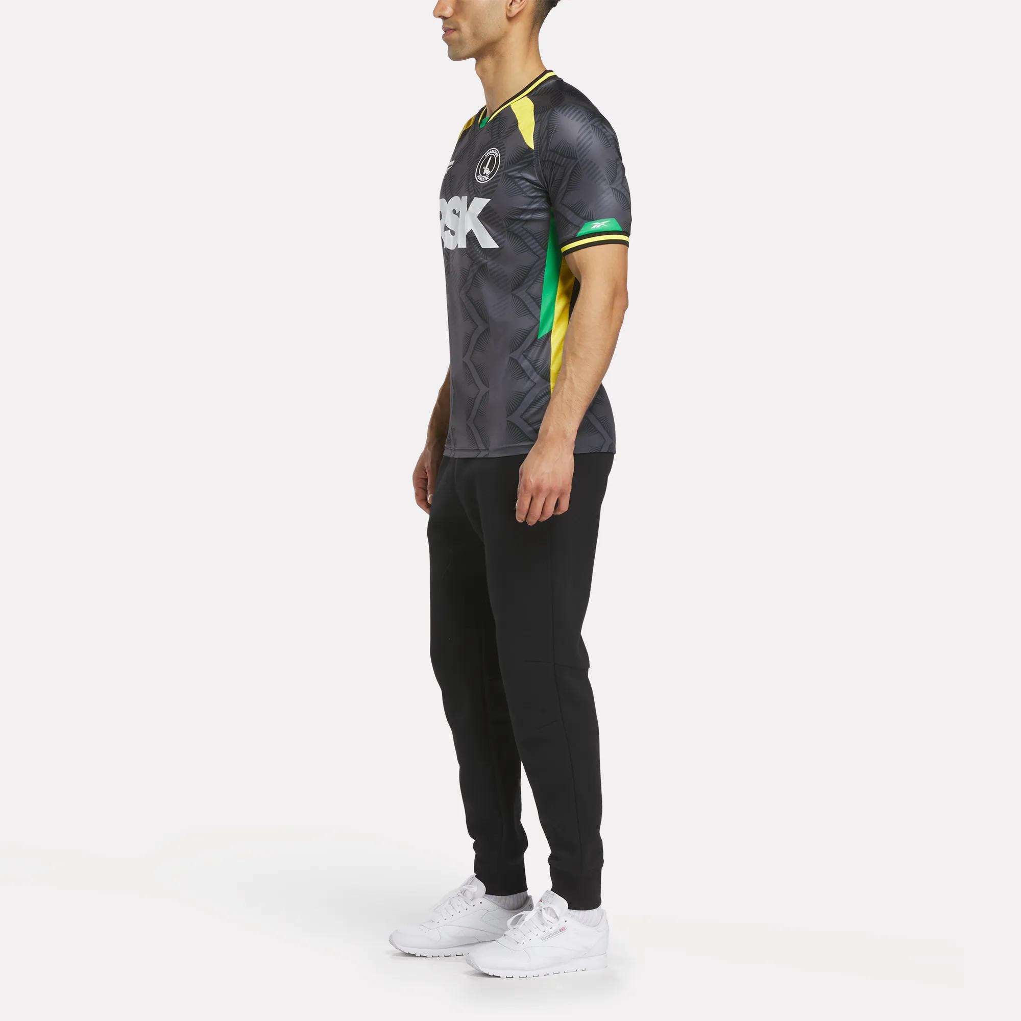 A man stands in profile against a white background, wearing the Reebok Men's Charlton Third Jersey with black pants and white sneakers. The shirt features white lettering plus green and yellow accents inspired by the team's third kit.