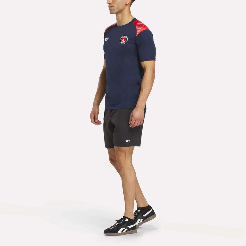 A man faces slightly left, wearing the Reebok Men's Charlton Training T-Shirt in navy blue with red accents, paired with black shorts and black sneakers with white soles. The shirt is made from breathable polyester for comfort.