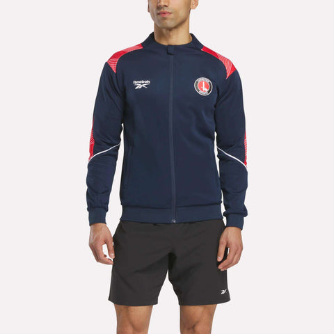 A man stands facing forward against a plain white background, wearing the Reebok Men's Charlton Training Jacket in navy blue with red and white accents, made from polyester doubleknit, paired with black shorts.