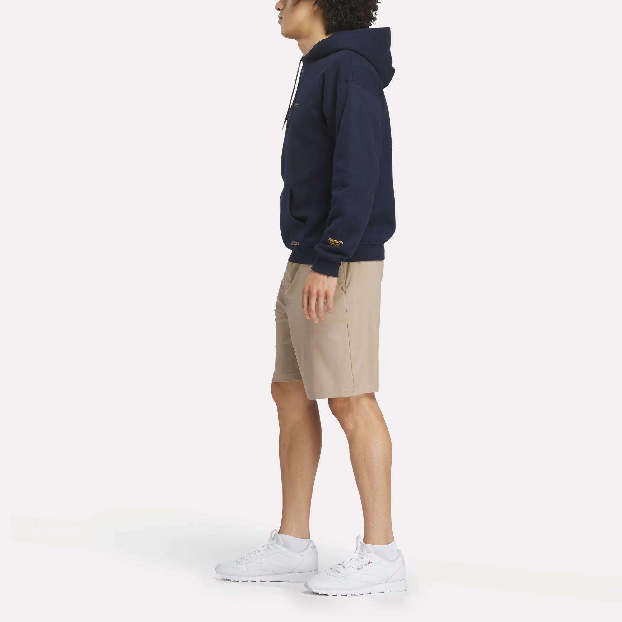 A person stands in profile wearing a Reebok Men's Crushers Hoodie in navy, beige shorts, and white sneakers against a plain white background.