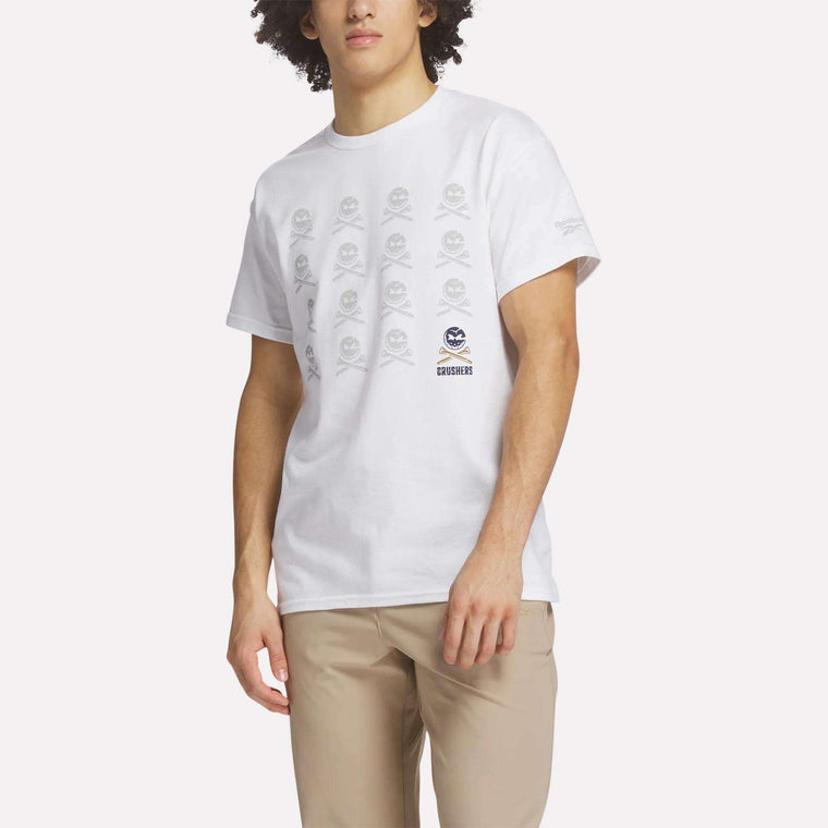 A man wears the Reebok Men's Crushers Icon Tee, featuring repeated gray skull and crossbones graphics on white, along with beige pants, standing against a plain light background.