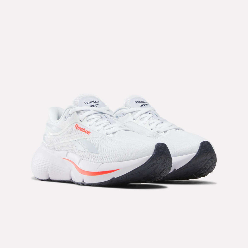 A pair of Reebok Women’s Zignition Running Shoes in white, featuring black soles and a subtle orange accent near the midsole, displayed on a plain white background. Engineered for comfort, these shoes provide springy cushioning that propels you forward with each stride.