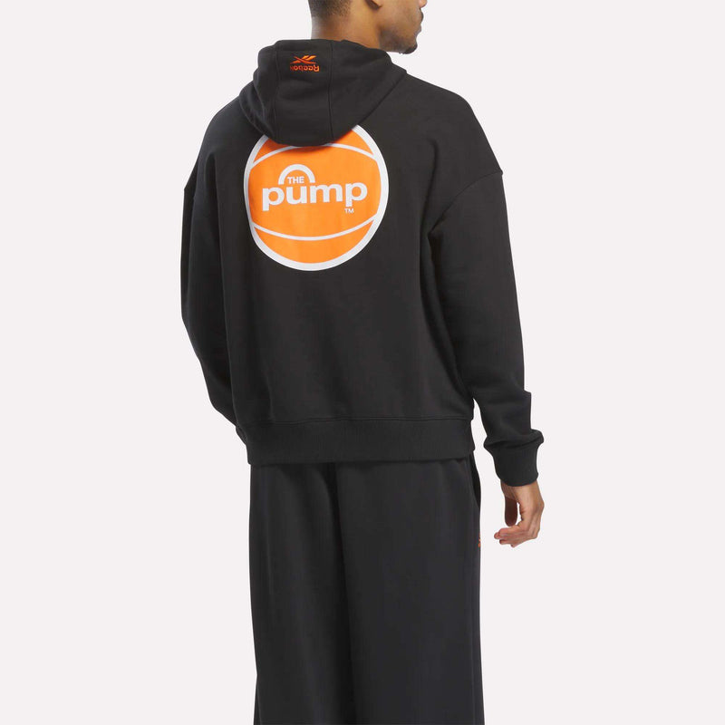 A person wearing a black Reebok Men's Pump OG Hoodie and pants, with the back of the hoodie showcasing a bold orange and white circular Pump logo inspired by retro basketball style.