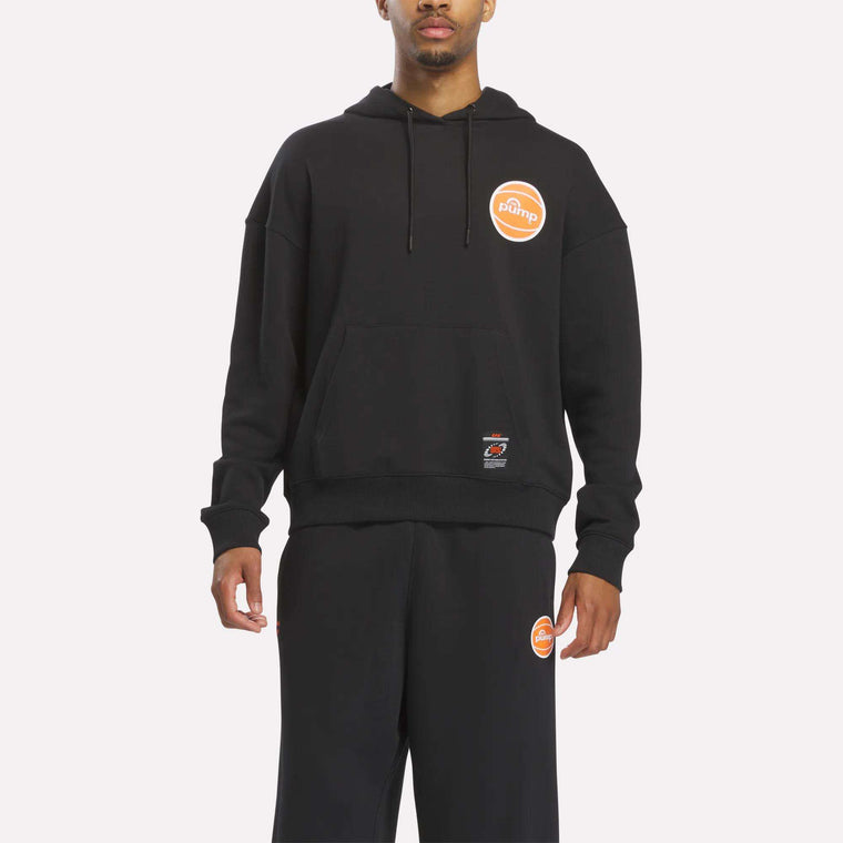 Dressed in the Reebok Men's Pump OG Hoodie in black paired with matching black sweatpants, both adorned with an orange circular Pump logo on the upper left side, a person channels retro basketball style while standing against a plain white background.
