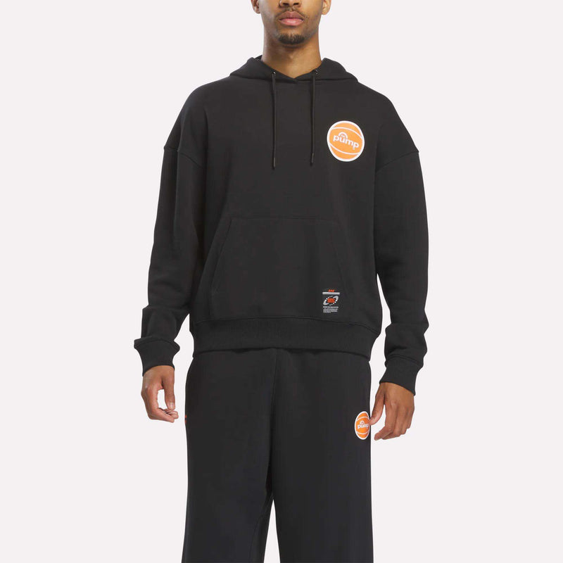 Dressed in the Reebok Men's Pump OG Hoodie in black paired with matching black sweatpants, both adorned with an orange circular Pump logo on the upper left side, a person channels retro basketball style while standing against a plain white background.