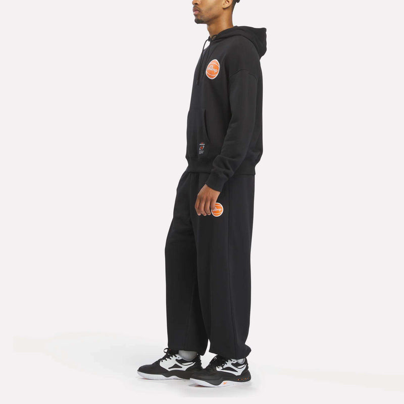 A person wearing a Reebok Men's Pump OG Hoodie in black stands sideways with their hands at their sides, paired with black sweatpants featuring orange patches and classic black and white sneakers inspired by retro basketball style, all set against a plain white background.