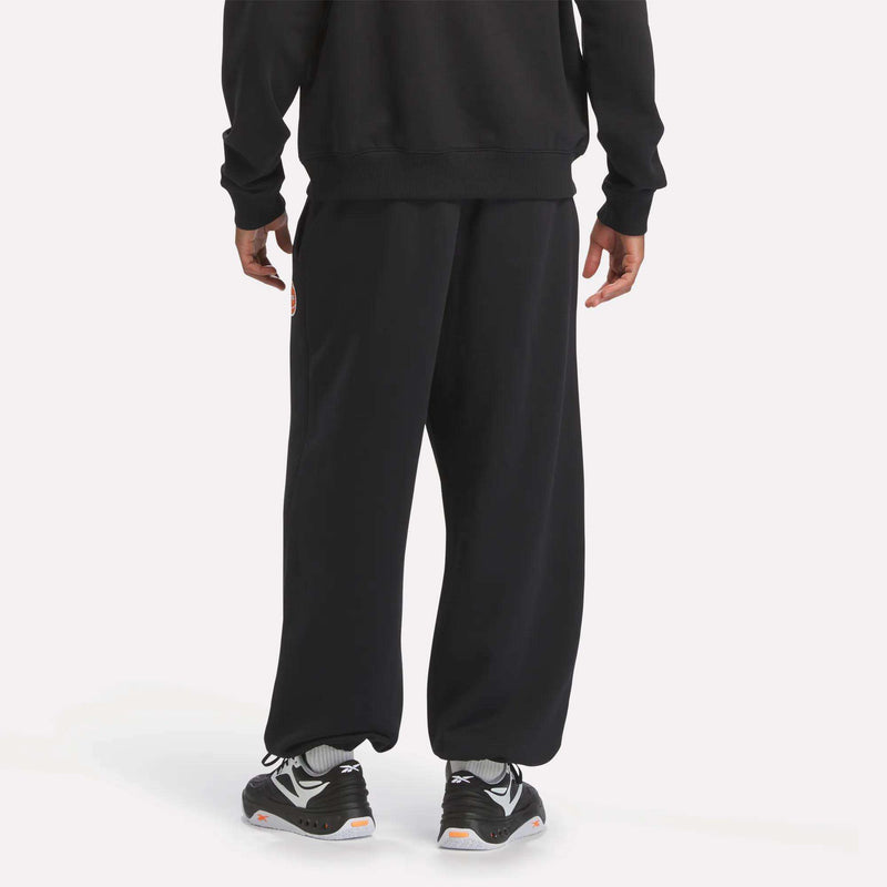 A person is wearing Reebok Men's Pump OG Joggers in black, paired with gray socks and black sneakers—possibly Reebok Pump—standing on a light background. The upper body and head are cropped out of the frame.
