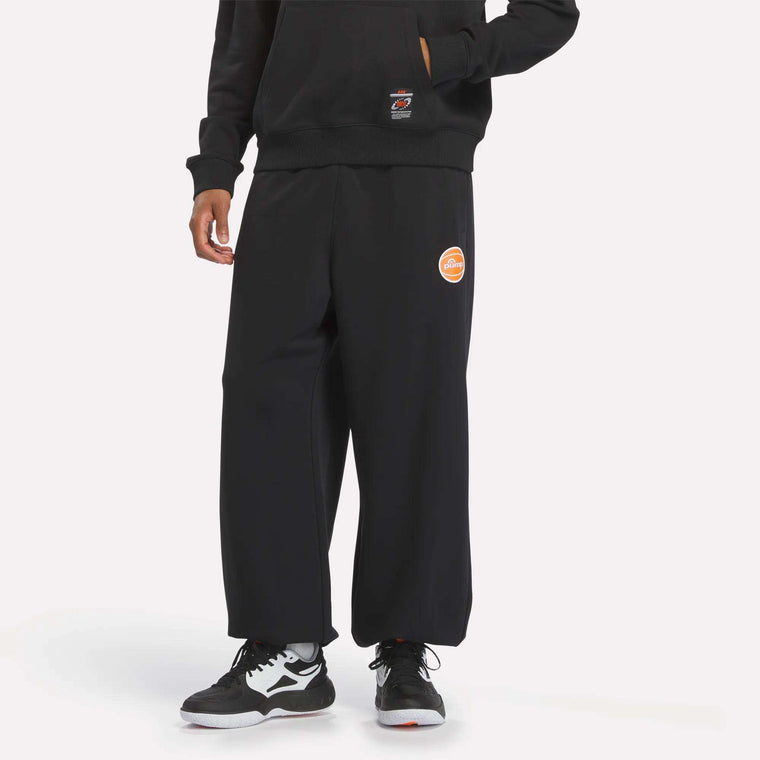 Person wearing loose black Men's Pump OG Joggers from Reebok, featuring a small logo detail, paired with a black hoodie with a subtle retro graphic and black-and-white Nike sneakers, standing against a plain white background.