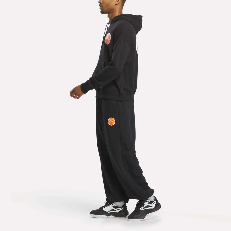 A person stands sideways against a white background wearing Reebok Men's Pump OG Joggers—black jogger pants featuring orange patches—paired with a matching black hoodie and black and white Reebok sneakers.