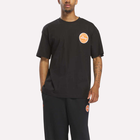 A person is wearing a loose-fitting black Reebok Men's Pump OG T-Shirt, which features an orange and white circular Pump logo, paired with black pants, standing against a plain light background.