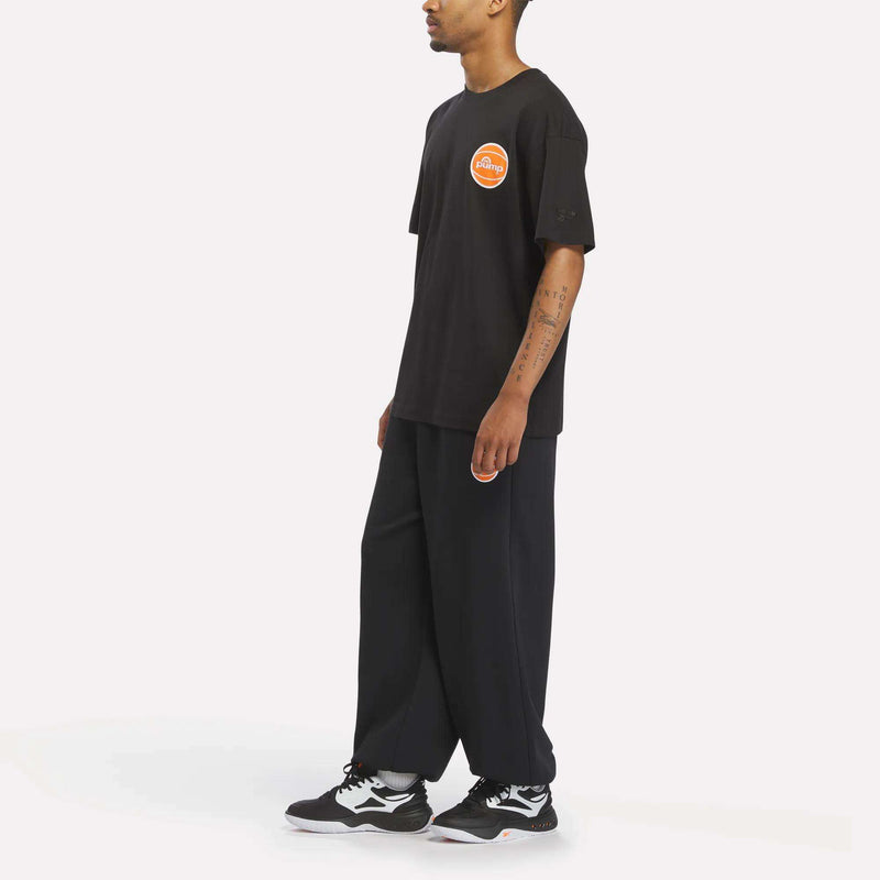 Man in a loose-fitting Reebok Men's Pump OG T-Shirt with a round orange logo, wide black pants, and Reebok Pump black-and-white sneakers standing sideways on a white background.