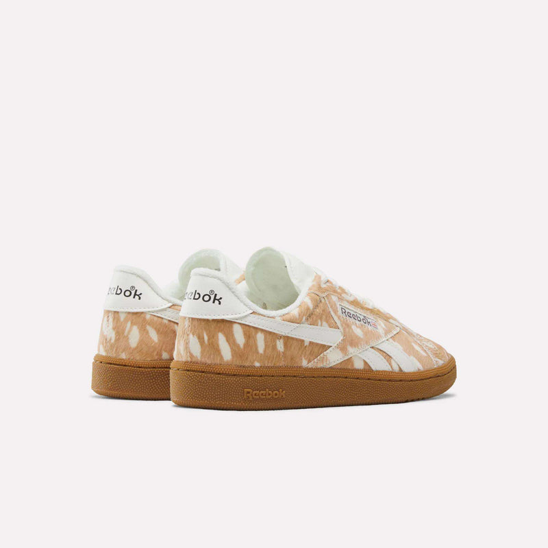 The Reebok Women’s Club C Grounds UK 1.0 Shoes showcase a bold animal print with a brown and white spotted fur pattern reminiscent of a fawn’s coat, complemented by gum soles. The Reebok logo is displayed on the side and heel, and the sneakers are set against a plain white background.