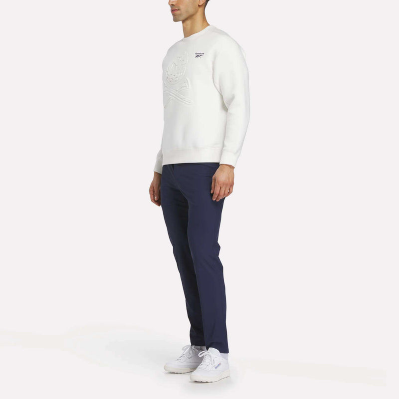 A man stands slightly to the side, wearing a white Reebok Men's Scuba Knit Crew Sweatshirt, navy blue pants, and white sneakers against a plain white background.