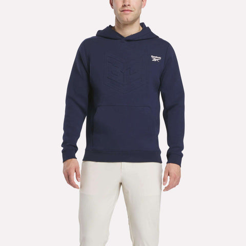 A man stands facing forward against a plain light background, wearing Reebok’s Men's Scuba Knit Hoodie Sweatshirt in navy blue with moisture-wicking fabric and an embossed chest design, paired with white pants.