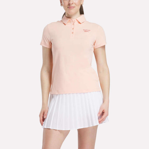 A woman in a Reebok Women's Micro Pique Sleeveless Polo Shirt in light pink and a white pleated skirt stands against a plain background. The polo shirt has UPF protection and a small logo on the left chest.