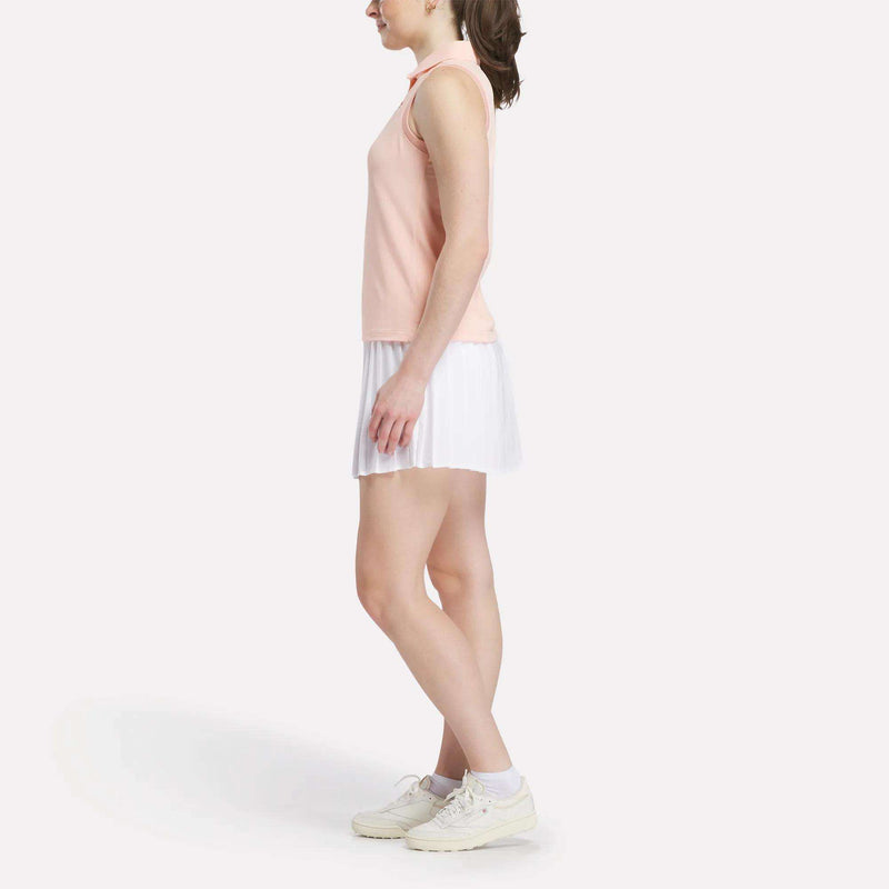 A woman in profile wears a Reebok Women's Micro Pique Sleeveless Polo Shirt in peach, paired with a white pleated skirt and white sneakers, set against a plain white background.