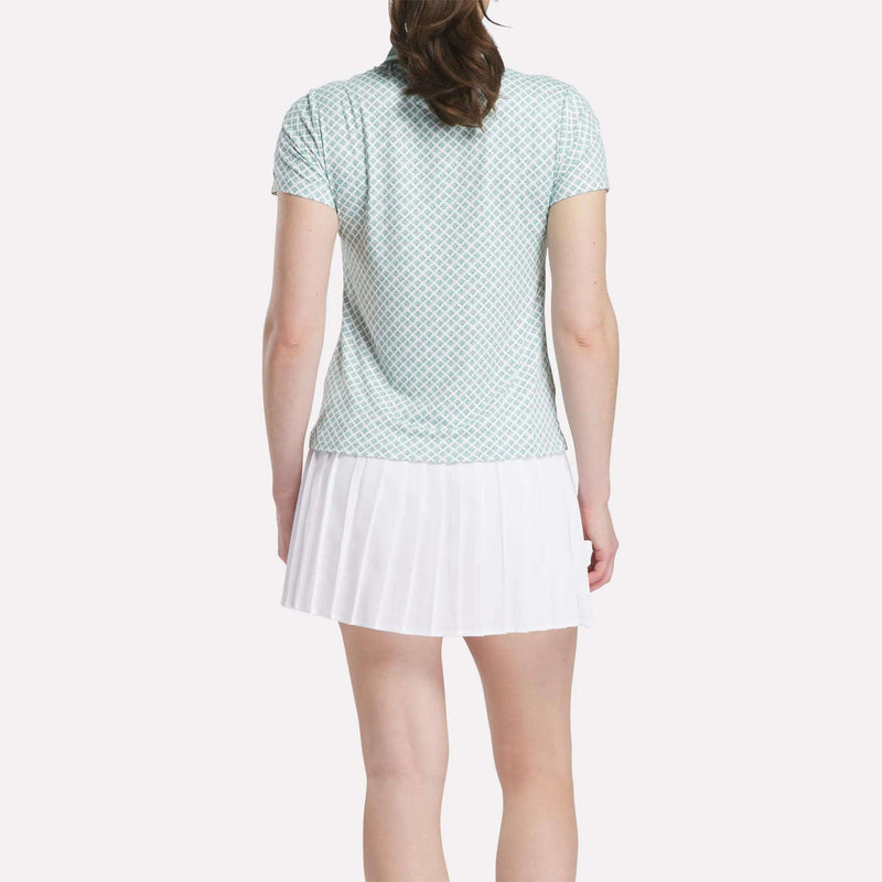 A woman with light skin and brown hair pulled back wears a Reebok Women's Foulard Print Sleeveless Polo Shirt and a white pleated skirt, standing against a plain white background that highlights her sporty style.