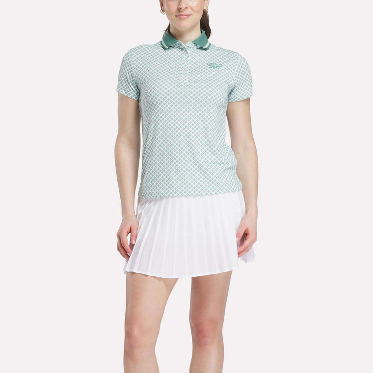 Woman wearing a Reebok Women's Foulard Print Sleeveless Polo Shirt and a white pleated skirt, standing against a plain white background.