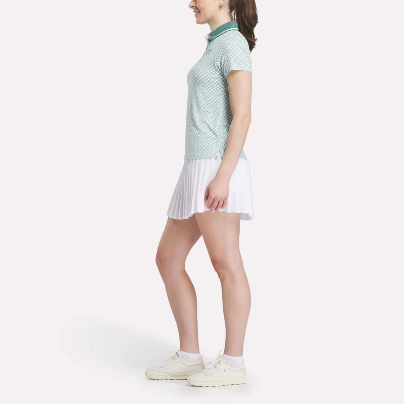A woman stands sideways in a light green Reebok Women's Foulard Print Sleeveless Polo Shirt, white pleated skirt, and white sneakers against a plain white background.