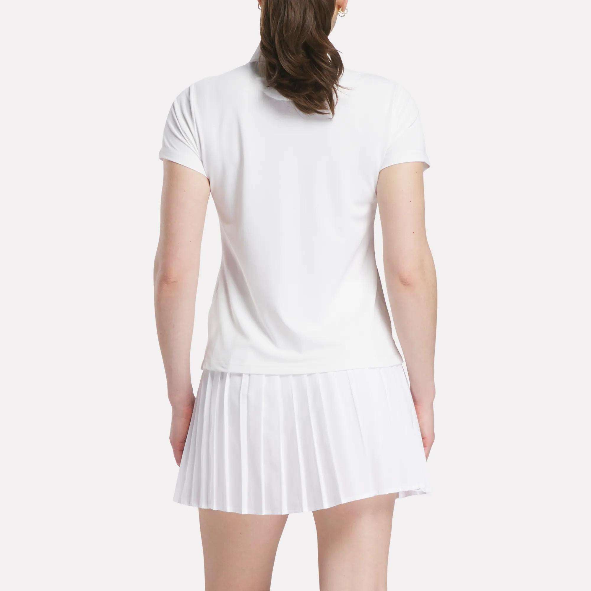 A woman is seen from the back wearing a Reebok Women's Micro Pique Polo Shirt with short sleeves and a white pleated skirt, standing against a plain light background.
