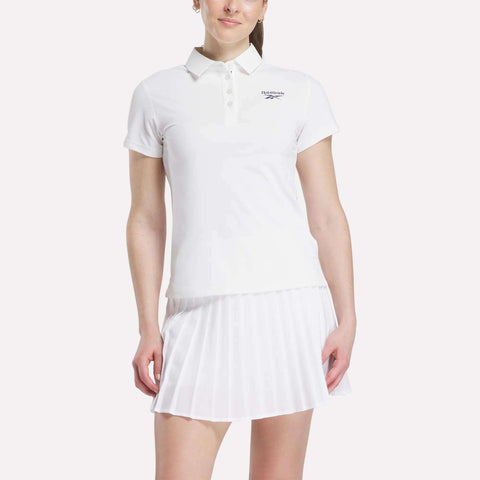 A woman in a white Reebok Women's Micro Pique Polo Shirt with a collar and short sleeves, paired with a white pleated skirt, stands against a plain light background.