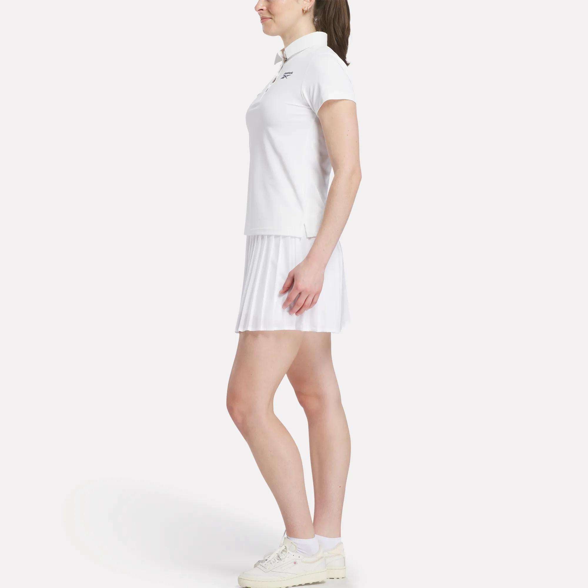 A woman stands in profile wearing a Reebok Women's Micro Pique Polo Shirt, a white pleated skirt, and white sneakers against a plain light background.
