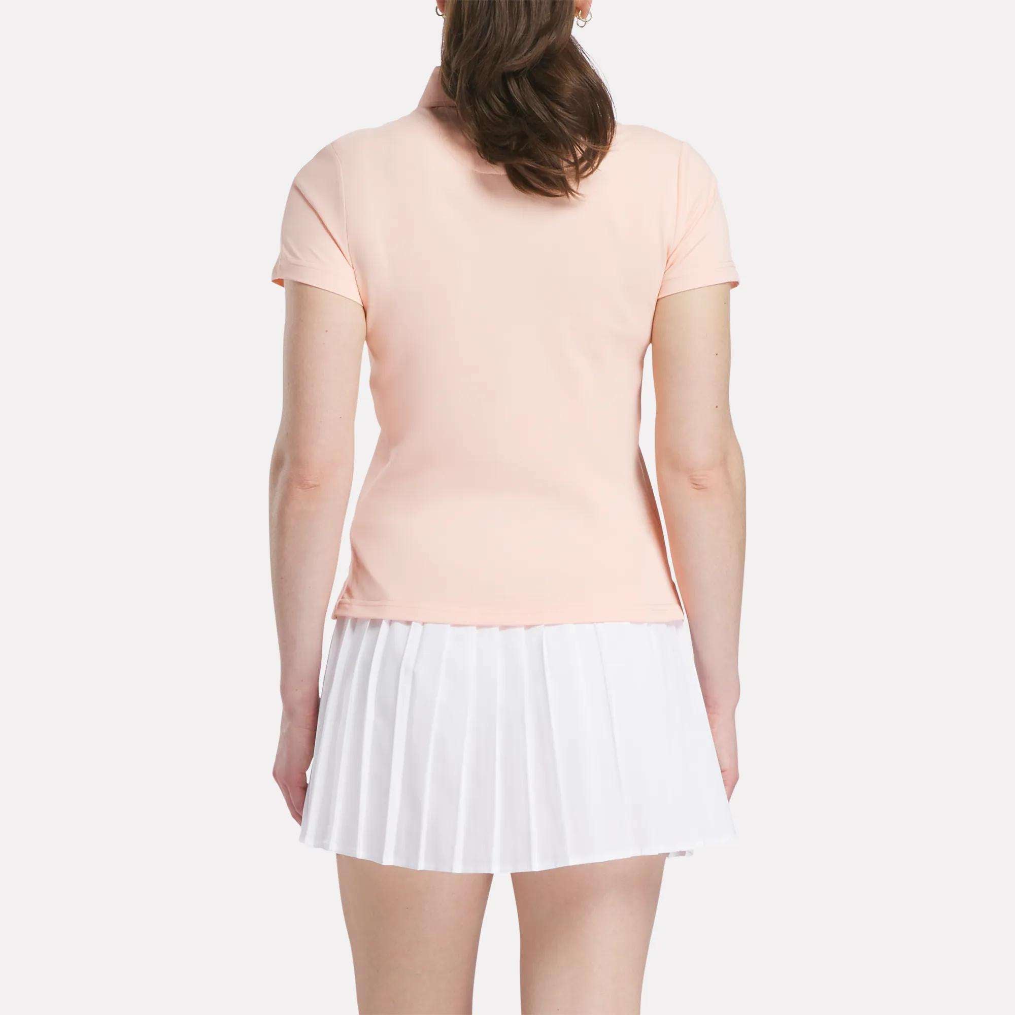 A woman faces away from the camera, dressed in a pale pink Reebok Women's Micro Pique Polo Shirt with stretch fabric and a white pleated skirt against a plain light background.