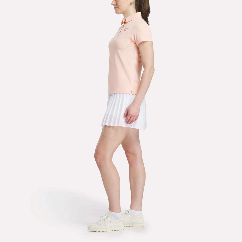 A woman stands in profile wearing a Reebok Women's Micro Pique Polo Shirt in light pink, paired with a white pleated skirt and white sneakers, all made from UPF-rated stretch fabric, posed against a plain white background.