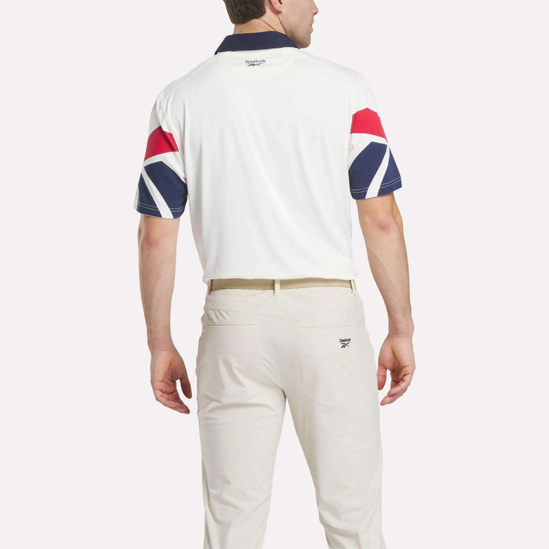 A man stands facing away, wearing the Reebok Men's Vector Sleeve Polo Shirt with UPF-rated stretch fabric, a white body, dark collar, red and blue geometric sleeve patterns, cream pants, all set against a plain white background.