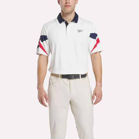 A man in a Reebok Men's Vector Sleeve Polo Shirt with moisture-wicking fabric, navy and red sleeve and collar accents, light-colored pants, and a black belt stands against a plain background.