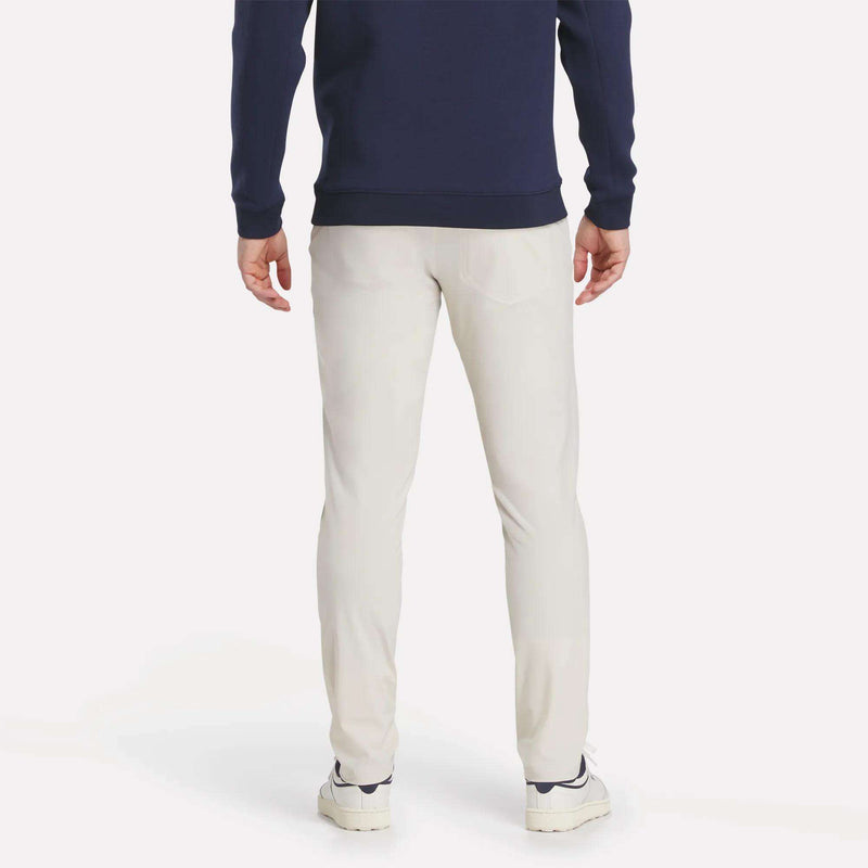 A person stands facing away, wearing a navy sweater, white sneakers, and Reebok Men's Favorite 5-Pocket Relaxed Fit Golf Pants in light beige against a plain white background.