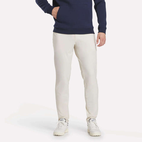 A person stands against a plain light background, showing only the lower half of their body, dressed in off-white Reebok Men's Favorite 5-Pocket Relaxed Fit Golf Pants paired with white sneakers and a navy hoodie.