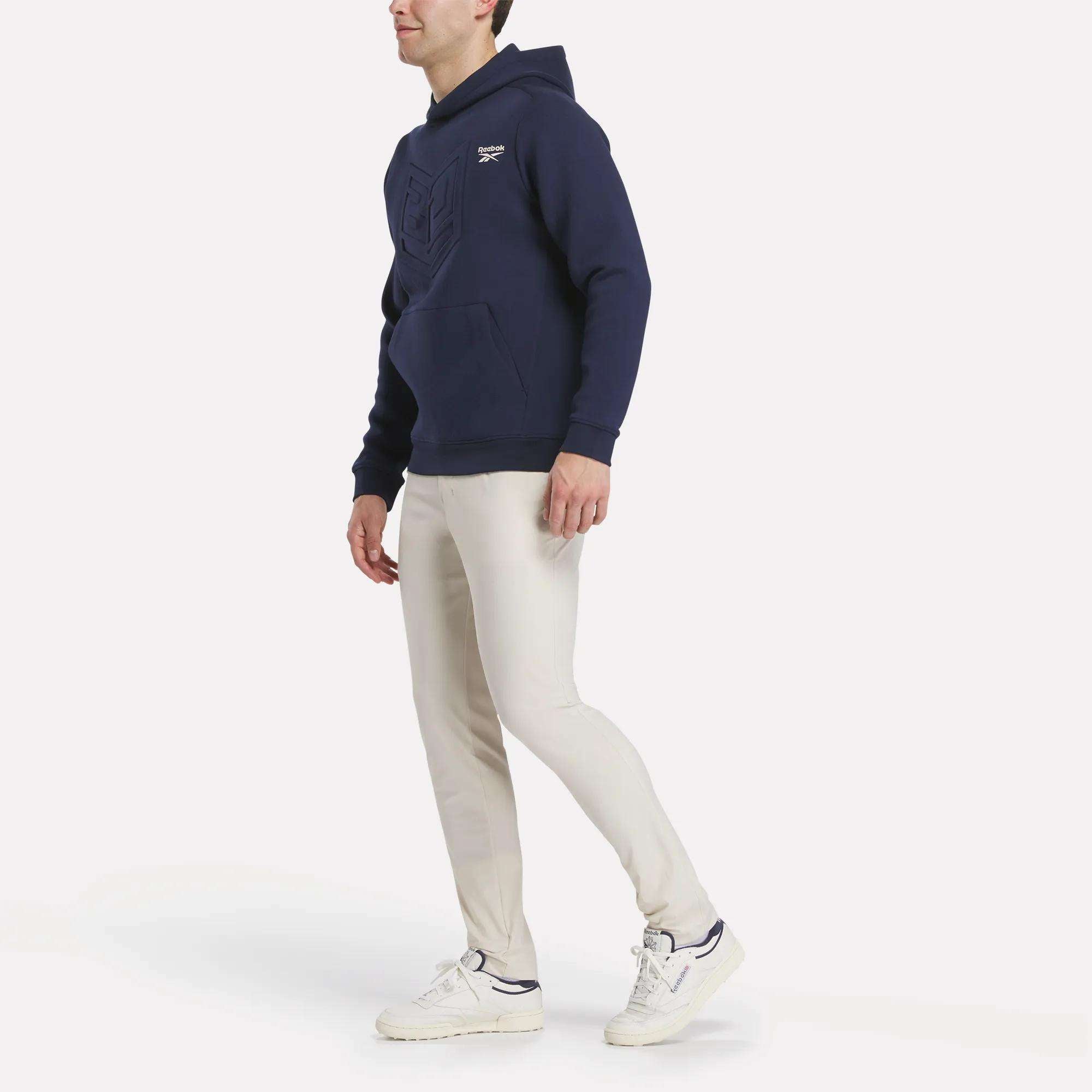 A person in a navy blue hoodie, Reebok Men's Favorite 5-Pocket Relaxed Fit Golf Pants made with RBK-DRY tech in light beige, and white sneakers stands against a white background. The hoodie has a front pocket and small chest logo.