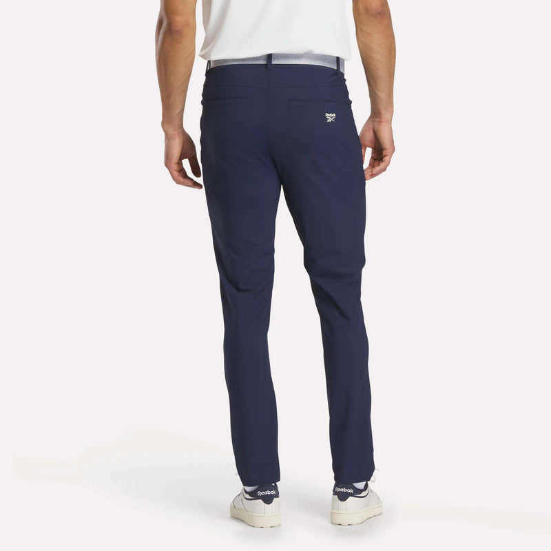 A man stands with his back turned, wearing Reebok Men's Favorite 5-Pocket Relaxed Fit Golf Pants in navy, paired with a white belt, white shirt, and white sneakers with dark soles against a plain white background.