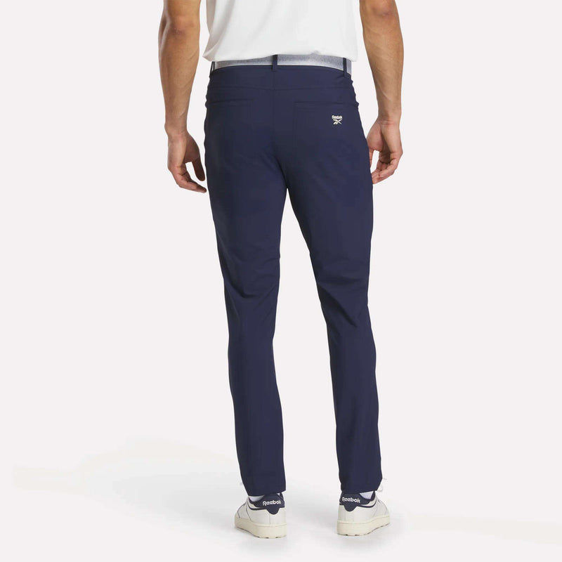 A person wearing Reebok Men's Favorite 5-Pocket Relaxed Fit Golf Pants in navy blue, paired with a white shirt and white sneakers, is standing with their back to the camera against a plain light background.