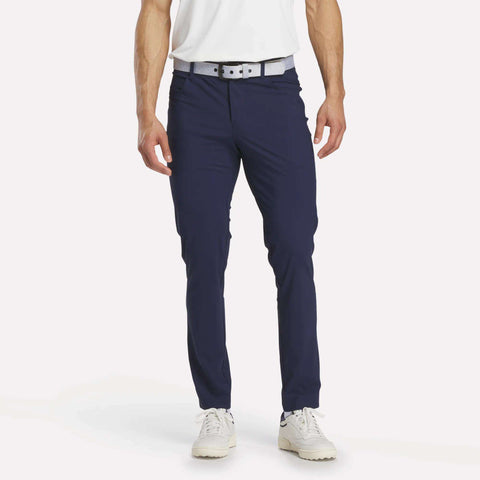 A person models Reebok Men's Favorite 5-Pocket Relaxed Fit Golf Pants in navy blue, paired with a white shirt, light gray belt, and white sneakers. The cropped photo highlights the sweat-wicking lower body against a plain white background.