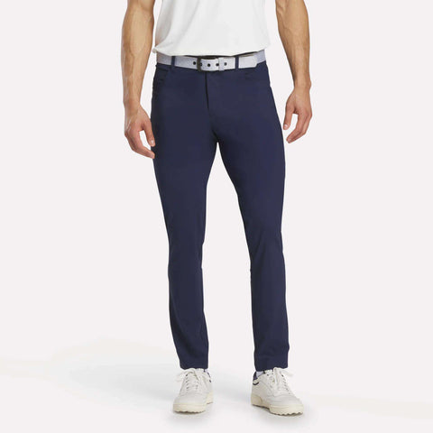 A man, head not visible, stands against a white background wearing Reebok Men's Favorite 5-Pocket Relaxed Fit Golf Pants in navy blue with a white belt, white shirt, and white sneakers. The wrinkle-resistant pants are the focal point.