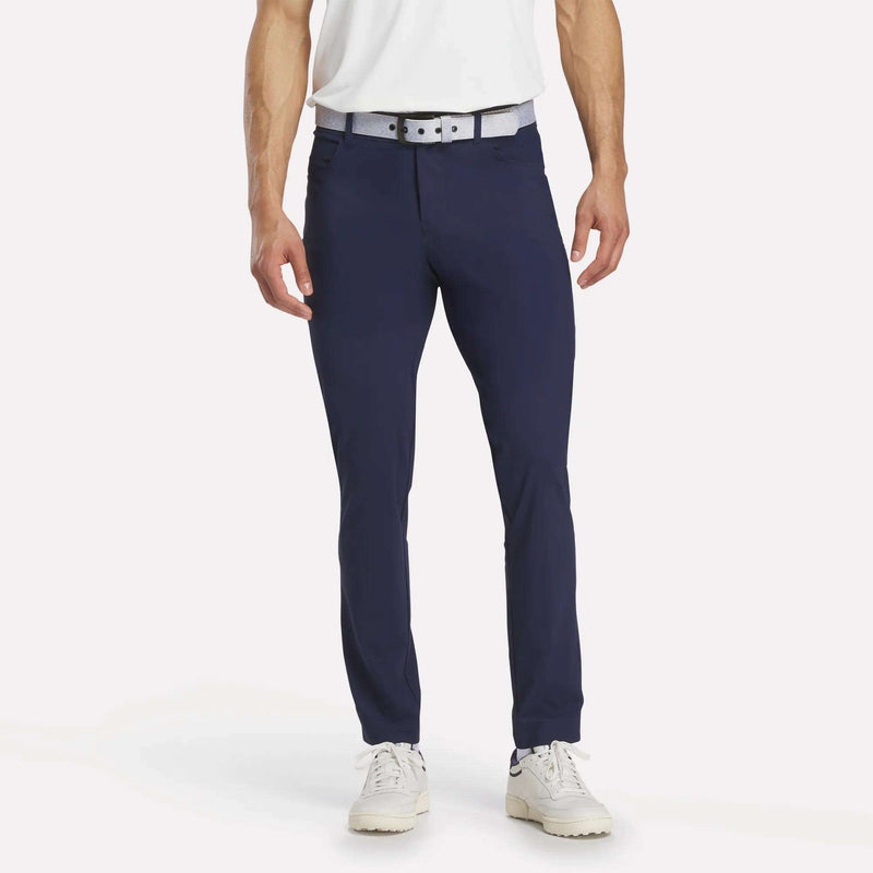 A person wearing a white shirt, white and gray belt, and white shoes stands against a plain white background, modeling Reebok Men's Favorite 5-Pocket Relaxed Fit Golf Pants in navy blue.