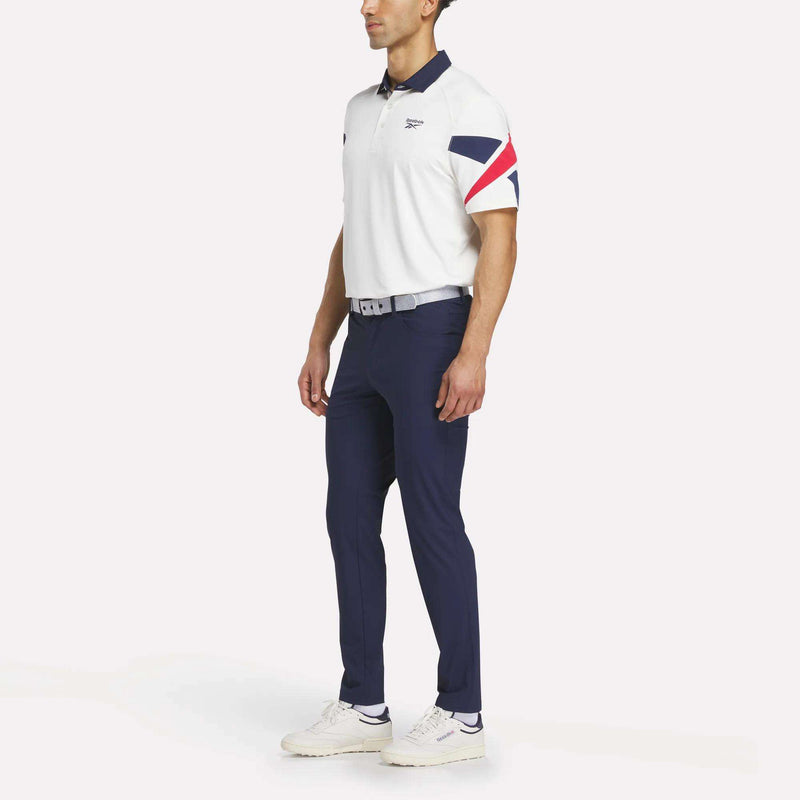 A man stands against a plain background wearing a white short-sleeve polo with red and navy sleeve stripes, Reebok Men's Favorite 5-Pocket Relaxed Fit Golf Pants in navy, a grey belt, and white sneakers.