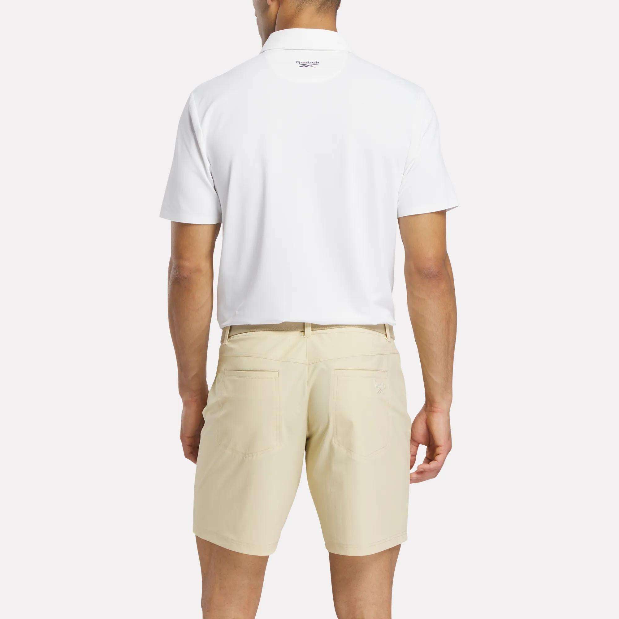 A man faces away from the camera, wearing a white Reebok Men's Bonded Pique Polo Shirt and light beige shorts against a plain background.