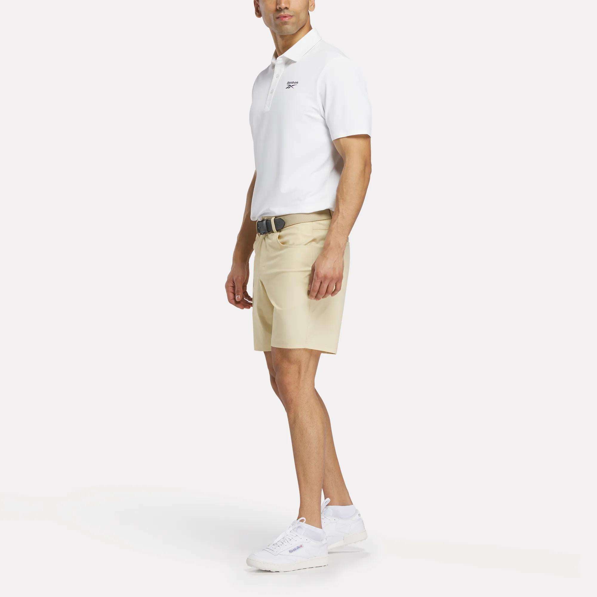 A man faces slightly left, wearing a white Reebok Men's Bonded Pique Polo Shirt, beige shorts, a black belt, and white sneakers against a plain white background.