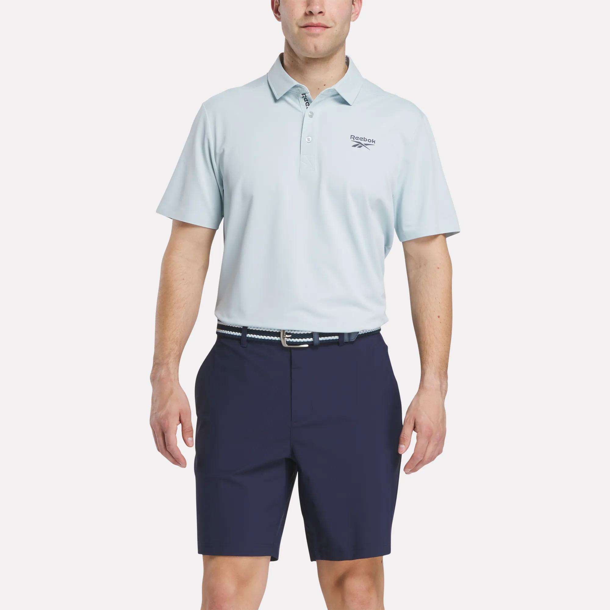 A man stands against a plain white background wearing a light blue Reebok Men's Bonded Pique Polo Shirt with a small left chest logo, navy shorts, and a patterned belt.