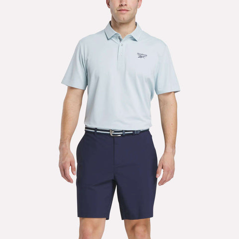 A man stands against a plain white background wearing a light blue Reebok Men's Bonded Pique Polo Shirt with a small left chest logo, navy shorts, and a patterned belt.