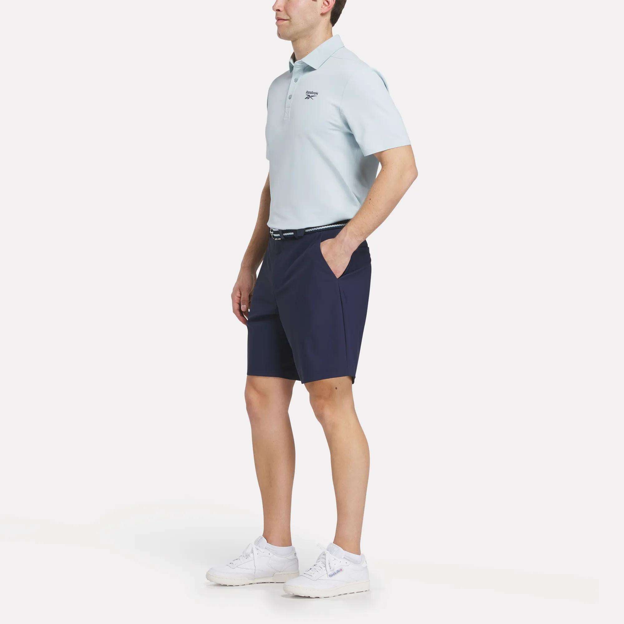 A man stands with hands in pockets, wearing a light blue Reebok Men's Bonded Pique Polo Shirt, navy shorts, a belt, and white sneakers against a plain white background.