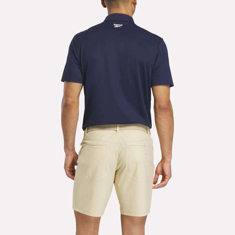 A man faces away from the camera, wearing a navy Reebok Men's Bonded Pique Polo Shirt and beige shorts. His arms rest at his sides against a plain, light background.