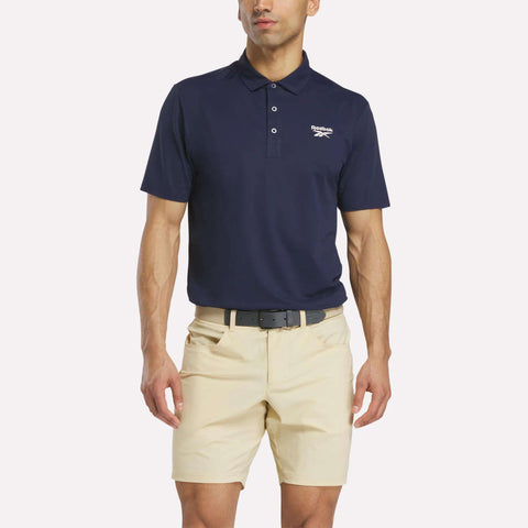 A man stands against a plain white background wearing a navy blue Men's Bonded Pique Polo Shirt by Reebok, featuring a white chest logo, paired with light beige shorts and a black belt.